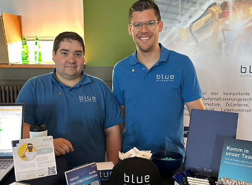 Two employees stand behind a blue automation trade fair stand